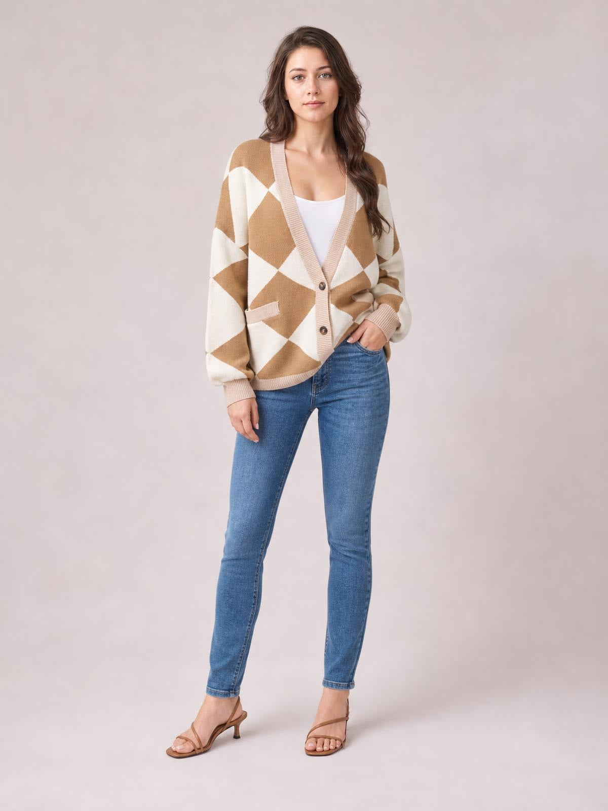 Model wears a tan and cream diamond pattern button-up knit cardigan with ribbed trim, front pockets, and a relaxed silhouette, posing with one hand in her jeans pocket.