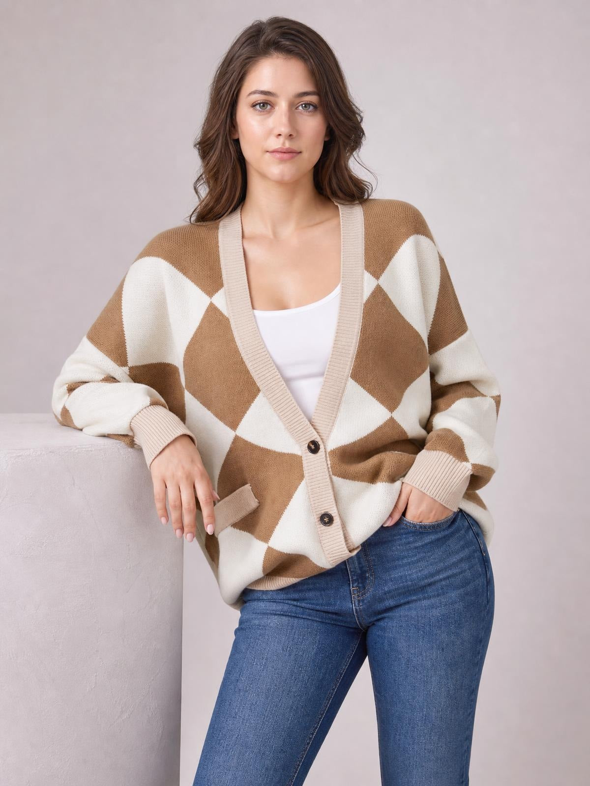 Model wearing a relaxed tan and cream diamond pattern button-up knit cardigan with ribbed trim, long sleeves, and front pockets, posing with one hand in her jeans pocket.