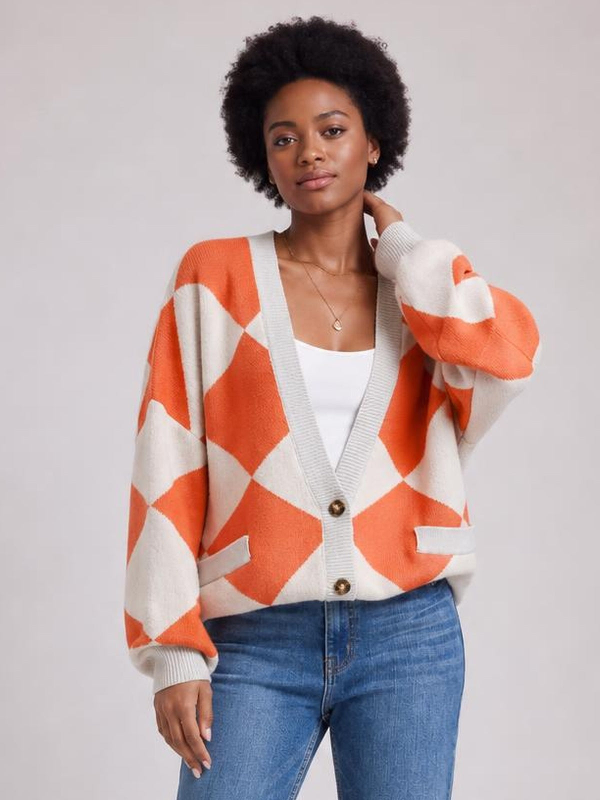 Model wearing an orange and cream diamond pattern button-up knit cardigan with a relaxed silhouette, ribbed trim, long sleeves, and front pockets.