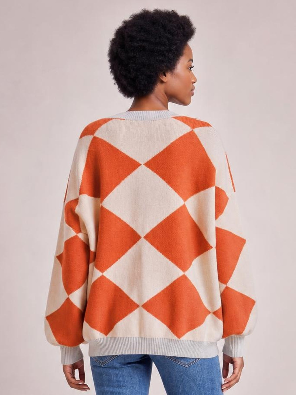 Model shown from behind wearing an oversized cream and orange diamond pattern knit cardigan with long sleeves, ribbed cuffs, collar, and hem.