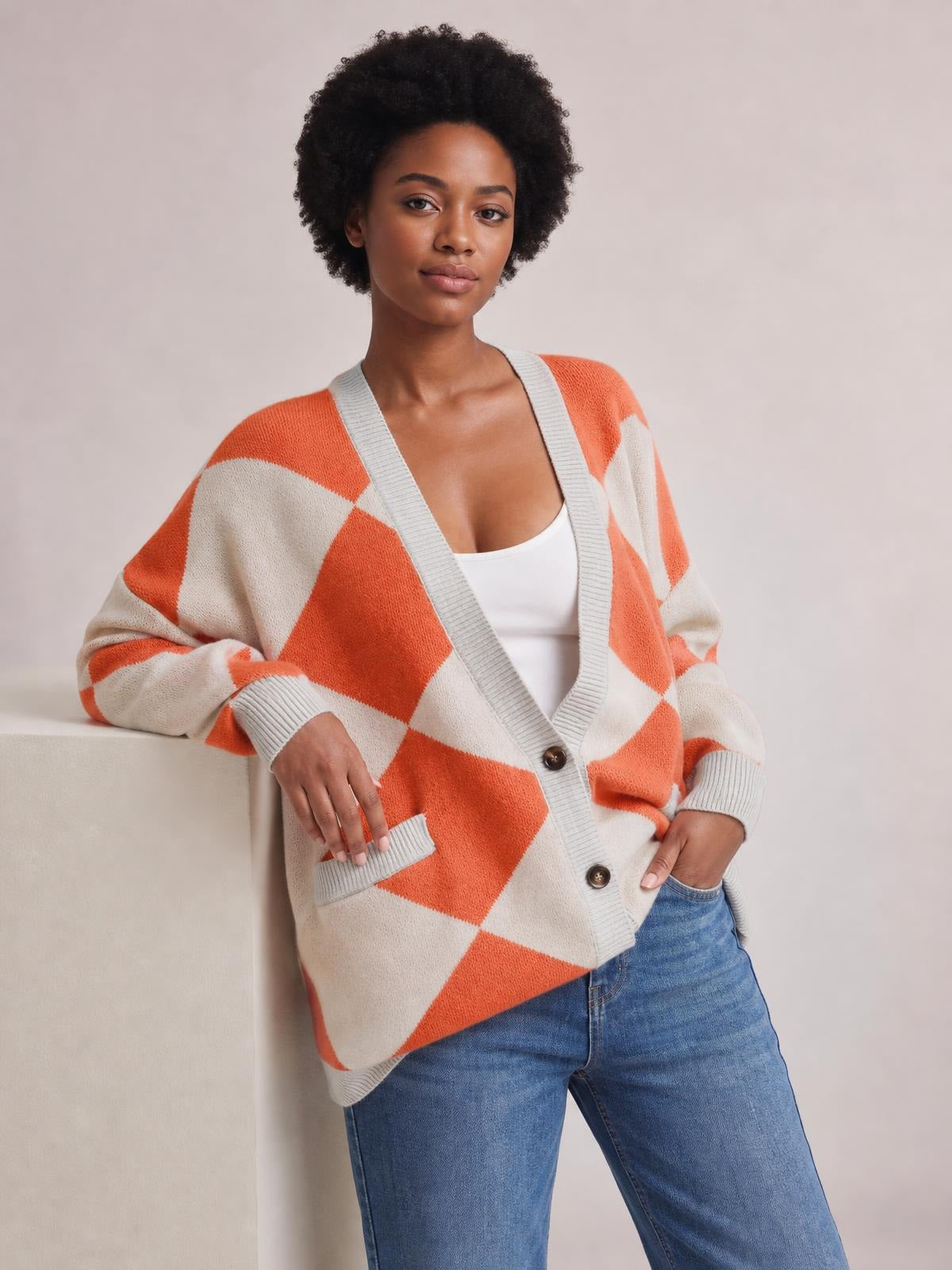 Model wears an orange and ivory diamond pattern knit cardigan with gray ribbed trim, button front, and pockets, posing with one hand in a pocket.
