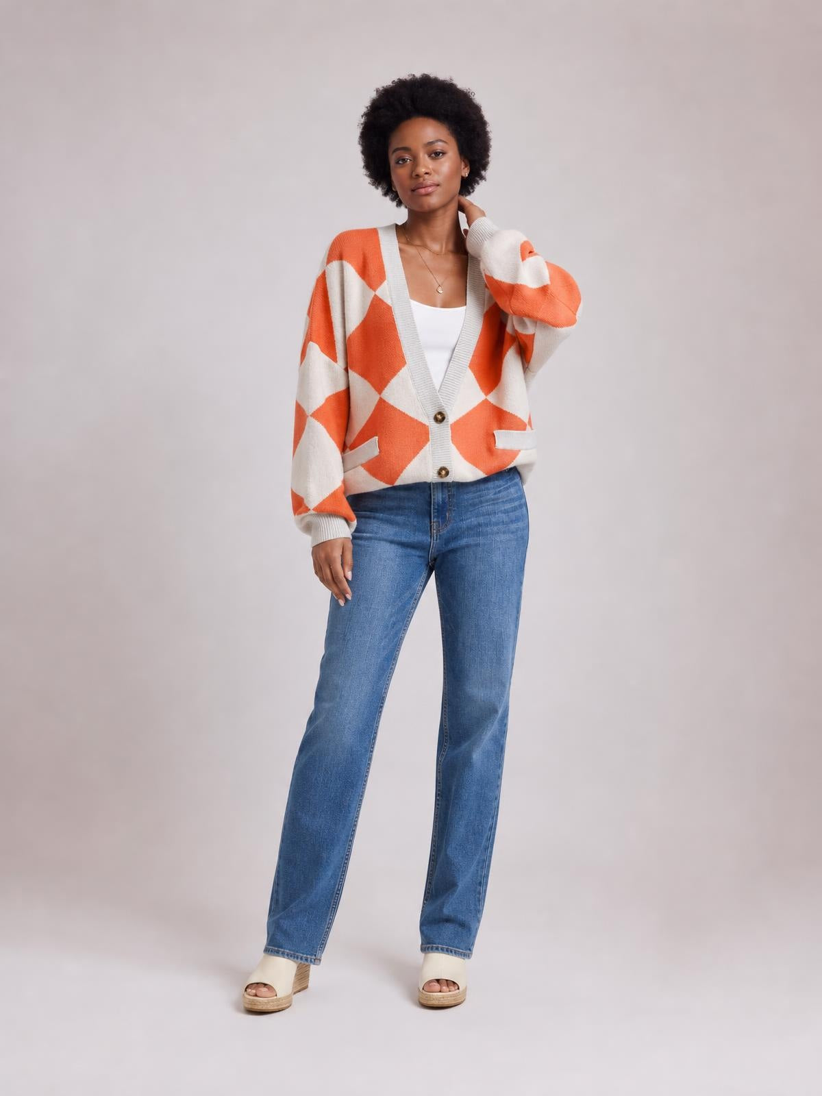 Model wearing an orange and cream diamond-pattern button-up knit cardigan with a relaxed silhouette, V-neck, ribbed trim, front buttons, and pockets.
