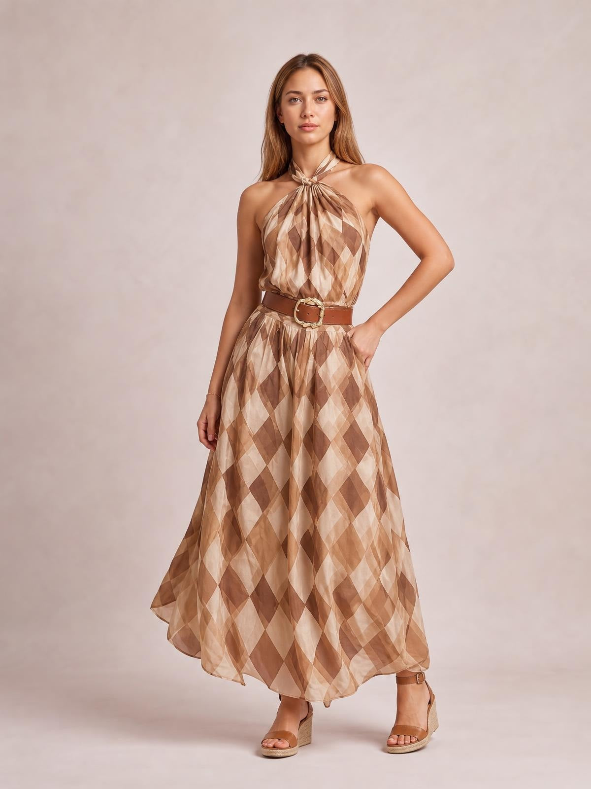 Model poses in a twisted halter neck maxi dress with a flowing A-line silhouette and brown, tan, and cream diamond pattern.