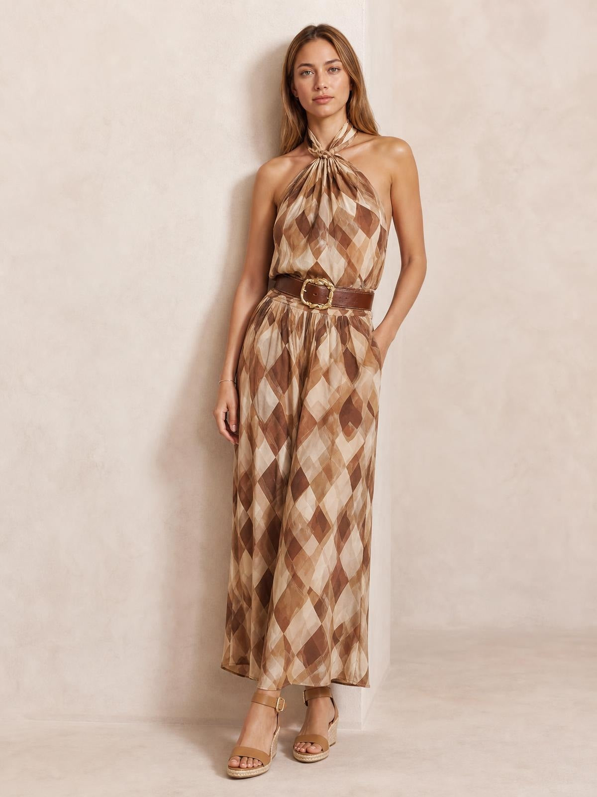 Model wearing a brown and beige diamond pattern halter neck maxi dress with a twisted neckline, flowing silhouette, and wide skirt, posing with one hand in a pocket.