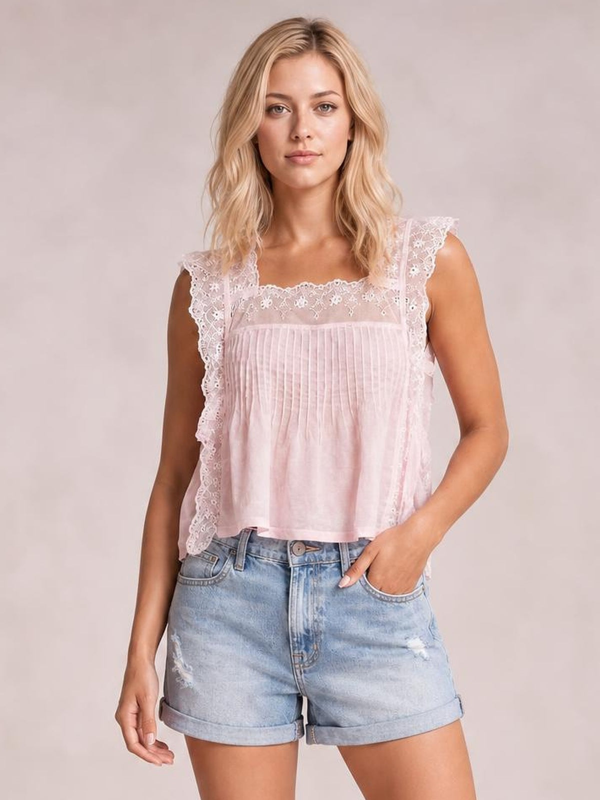 A woman modeling a light pink sleeveless top with delicate lace trim and ruffle details, paired with light-wash denim shorts, against a solid light beige studio background.