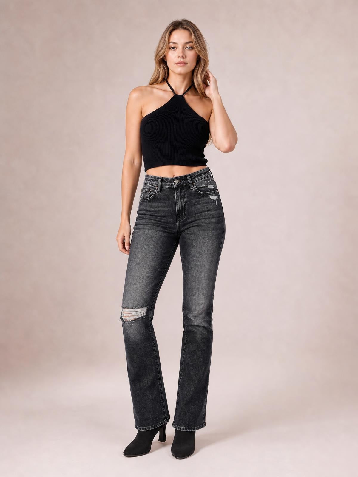 Standing with one hand at her hair, a model wears dark gray distressed flare jeans with a high waist, button closure, front pockets, and a ripped knee.