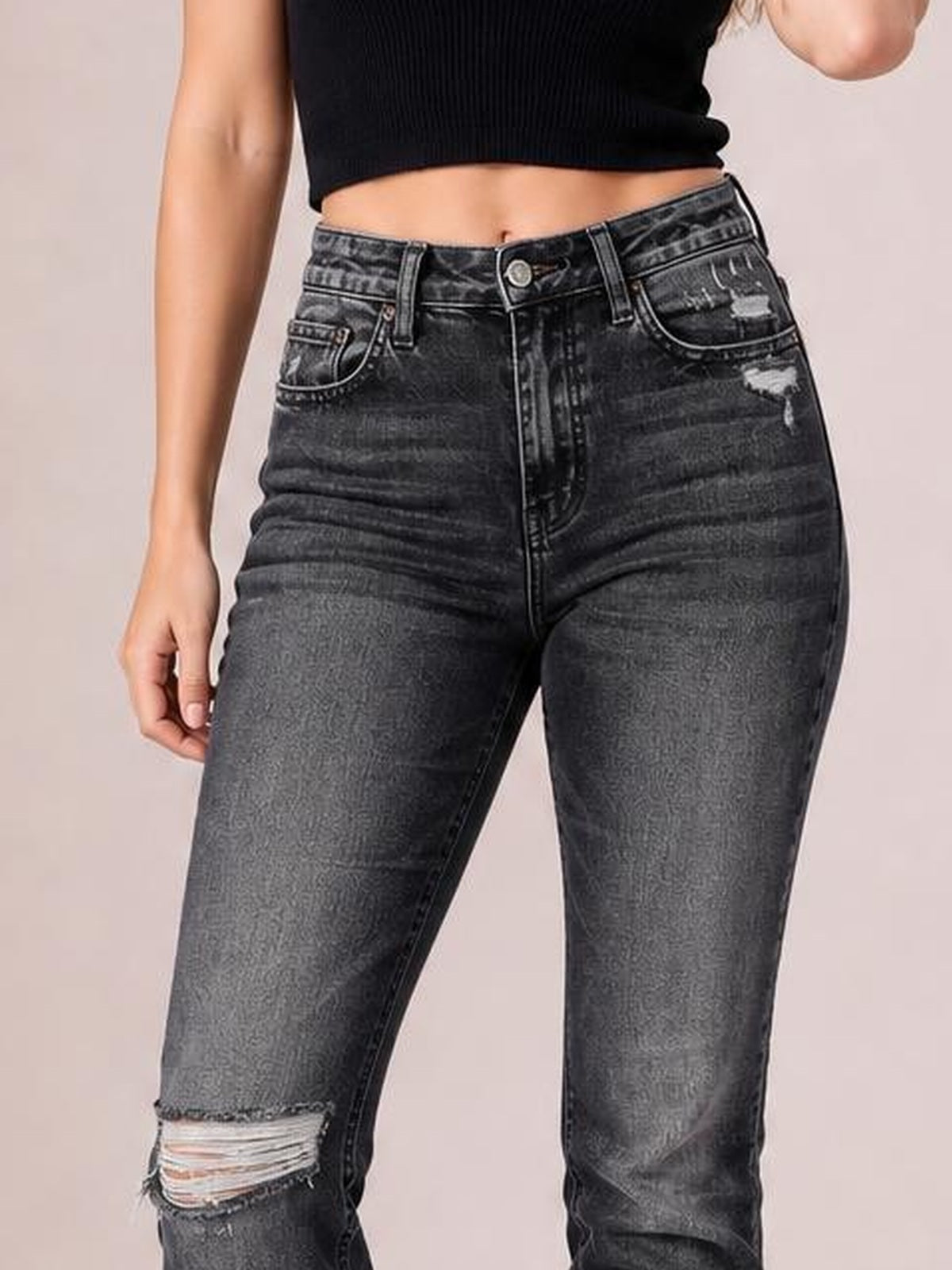 A model wears dark gray distressed jeans with a button closure, belt loops, front pockets, and a fitted high-waisted silhouette, posing with one hand at her side.