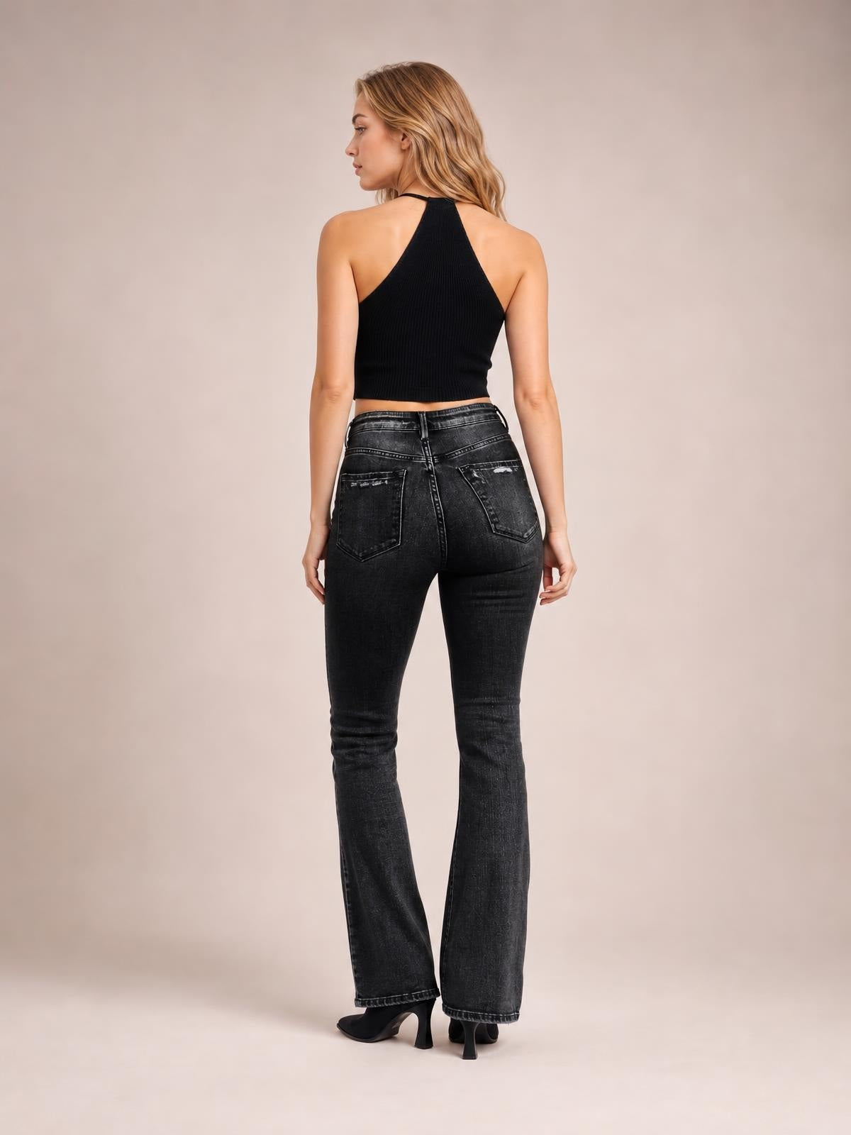 Model stands facing away in dark gray distressed flare jeans with a fitted high waist, button closure, and two back patch pockets.