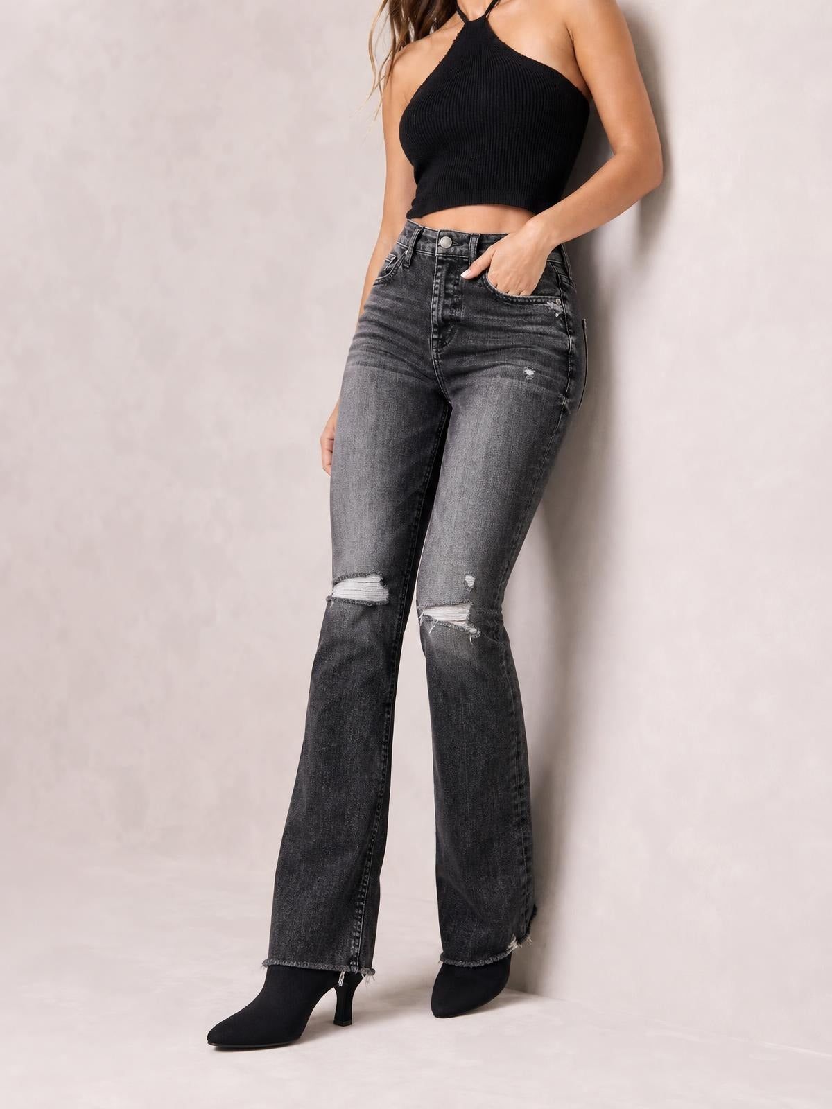 Model poses in distressed charcoal gray flare jeans with a button closure, front pockets, fitted hips, ripped knees, and raw hems.