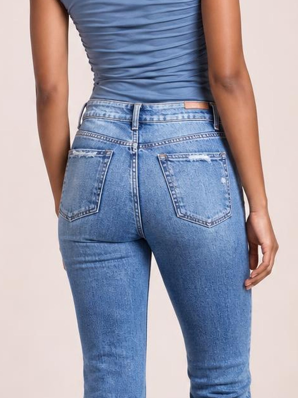 A model wears light blue distressed jeans in a back-facing pose, featuring a fitted silhouette, belt loops, contrast stitching, and two pocketed back pockets.