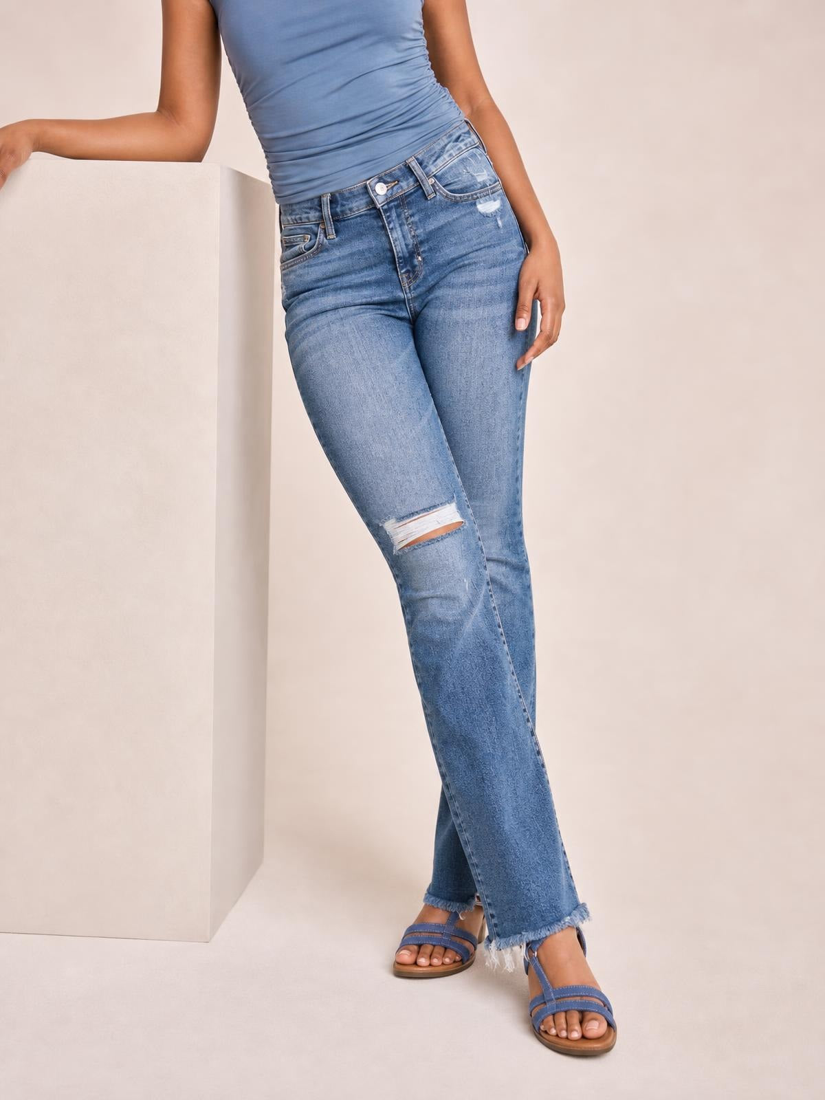 Model poses in light blue distressed flare jeans with a button closure, belt loops, front pockets, a ripped knee, and frayed hems.
