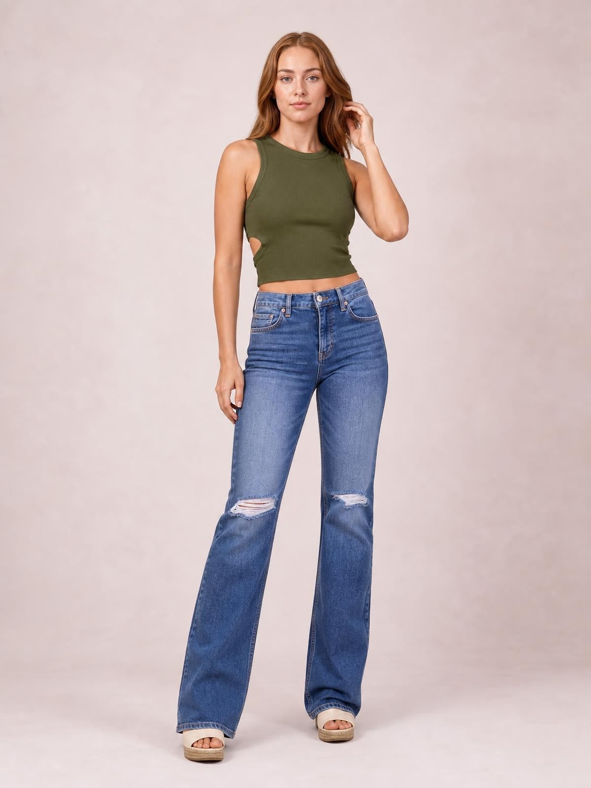 Model stands in blue distressed flare jeans with a button fly, front pockets, ripped knees, and a high-rise silhouette, gently touching her hair.