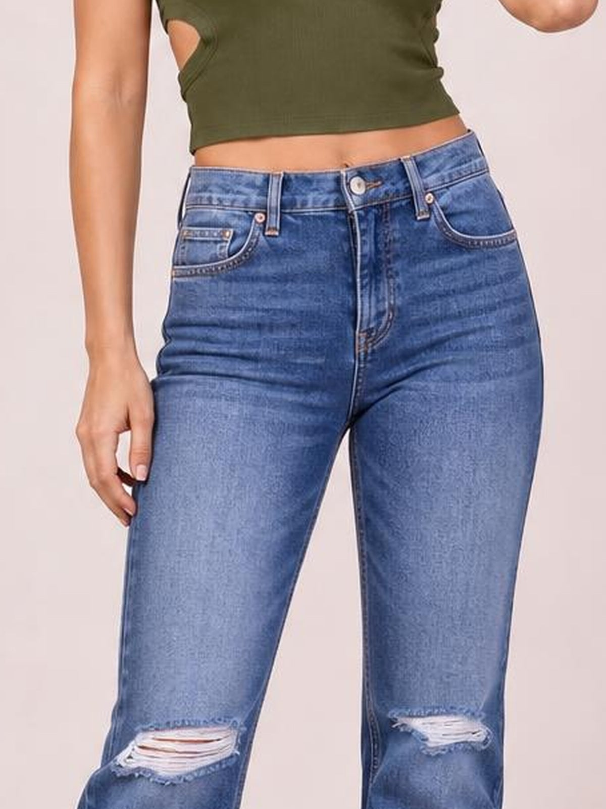 A model wears blue distressed jeans with a button fly, belt loops, front pockets, and ripped knees, standing with one hand resting on her thigh.