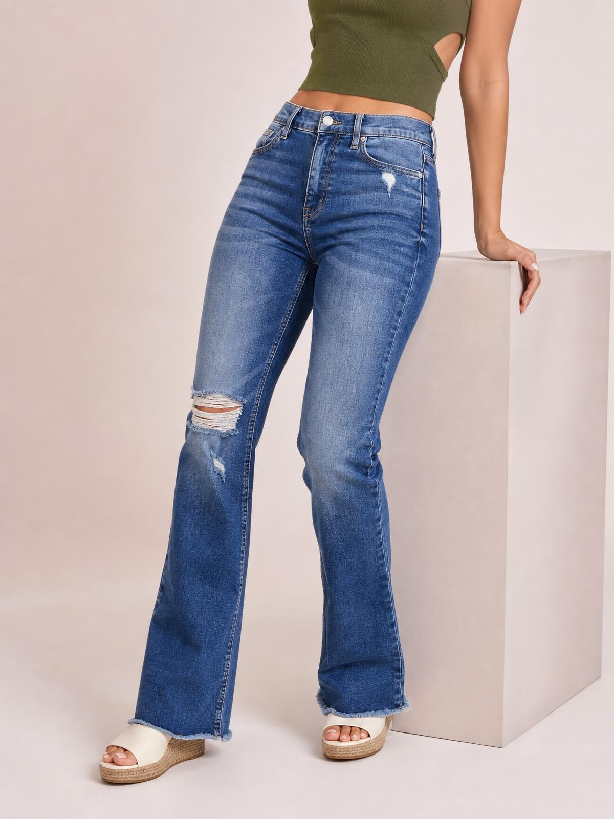 A model wears blue distressed flare jeans with a buttoned waistband, front pockets, ripped knees, a fitted silhouette, and frayed flared hems.