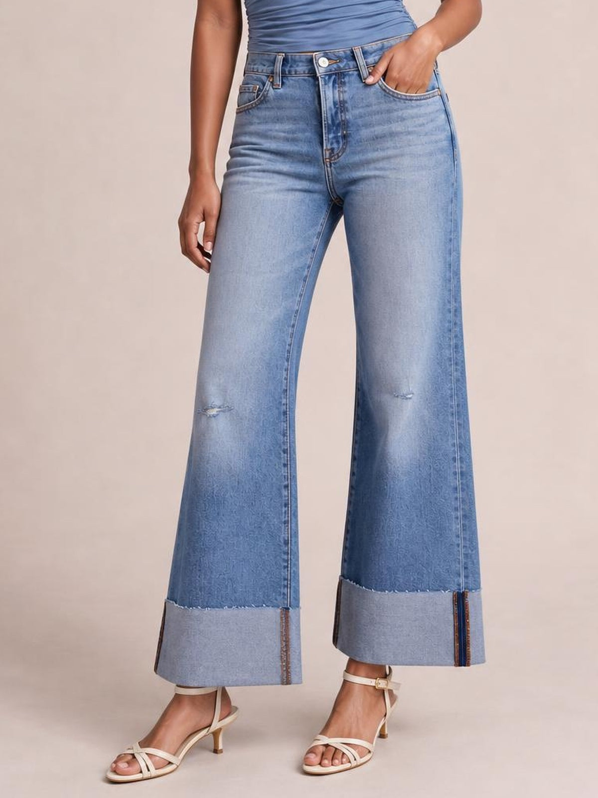 A model wears light-blue distressed high-waisted flare jeans with a wide-leg silhouette, small knee abrasions, raw hems, and one hand in a pocket.