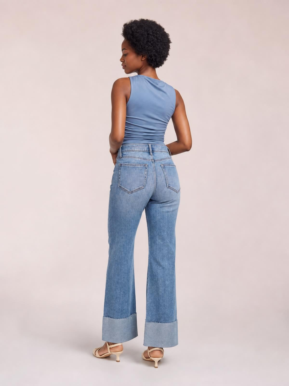 A model stands turned away wearing light-wash distressed high-waisted flare jeans with back pockets, a fitted seat, and raw hems.