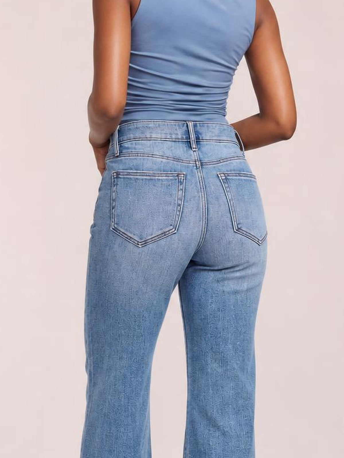 Model poses with hands in her pockets, showing the back of light blue distressed high-waisted flare jeans with contrast stitching and back patch pockets.
