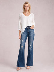 Model poses with one hand in the pocket, wearing dark blue distressed high-waisted flare jeans with a fitted waist, pocketed construction, and wide raw hems.
