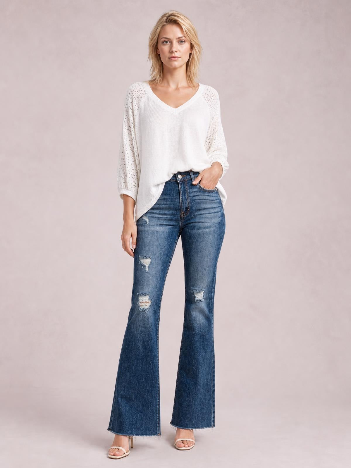 Model poses with one hand in the pocket, wearing dark blue distressed high-waisted flare jeans with a fitted waist, pocketed construction, and wide raw hems.