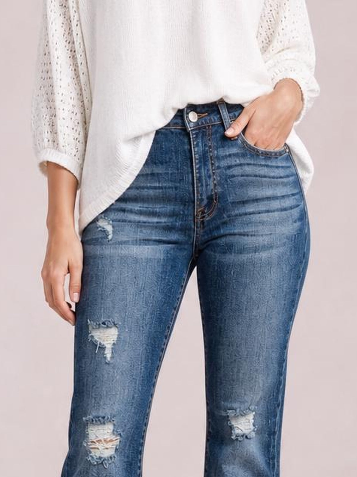 A model wears distressed high-waisted blue jeans with front pockets, a fitted waist and thighs, and ripped details at the left thigh and knees.