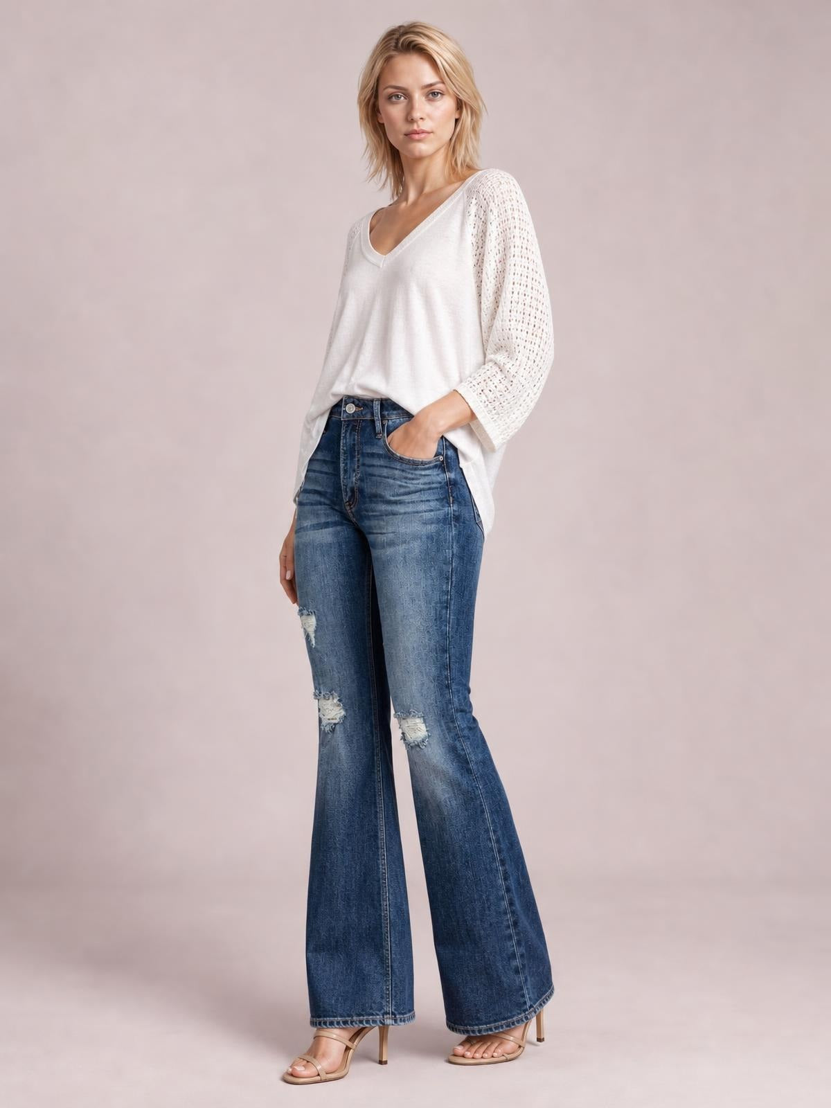 A model poses with one hand in the pocket of blue distressed high-waisted flare jeans, showing front pockets, ripped knees, and a wide-leg silhouette.