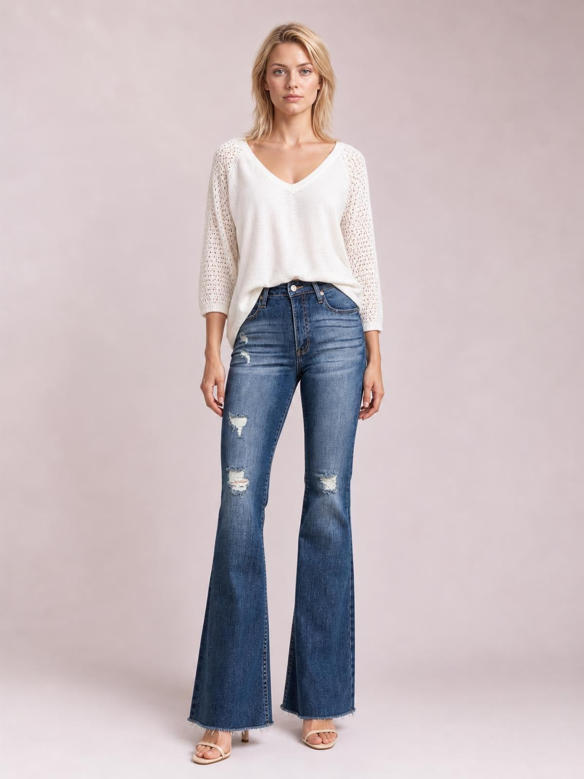 Model stands facing forward in blue distressed high-waisted flare jeans with front pockets, fitted thighs, ripped knees, and wide raw hems.