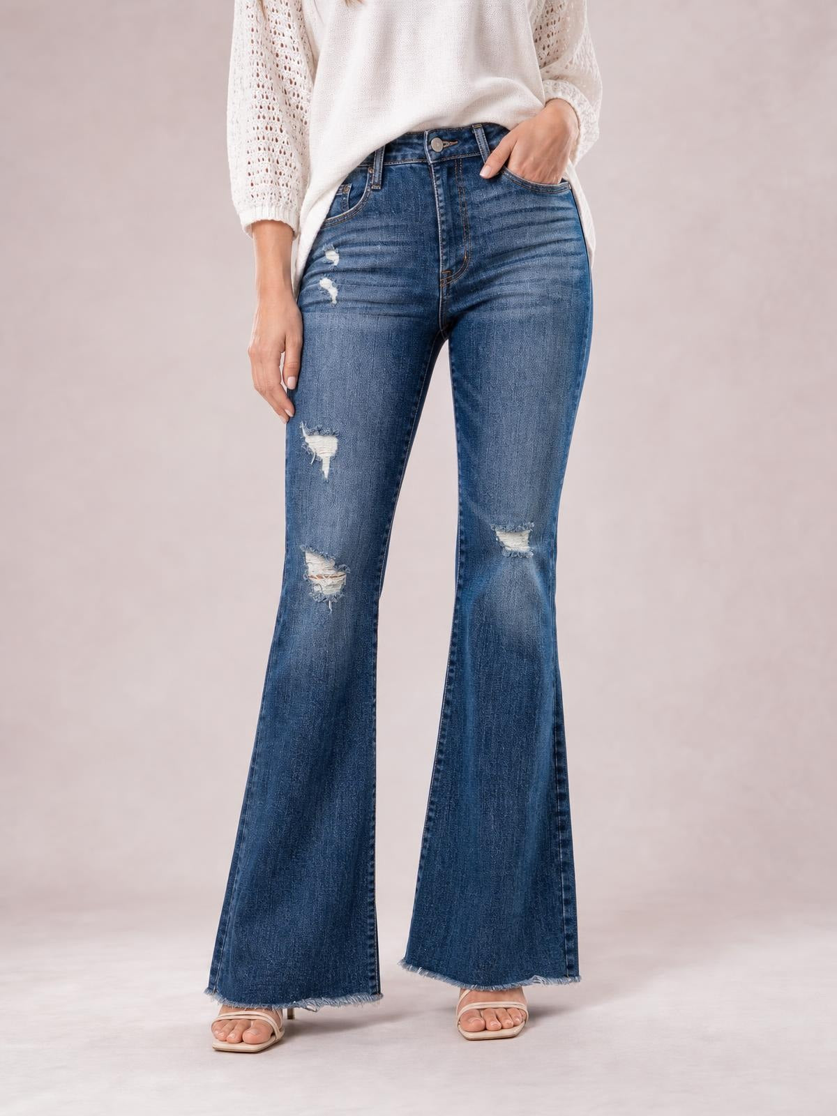 Model wears dark blue distressed high-waisted flare jeans with front pockets, whiskering, ripped knees, and frayed hems while standing with one hand in a pocket.