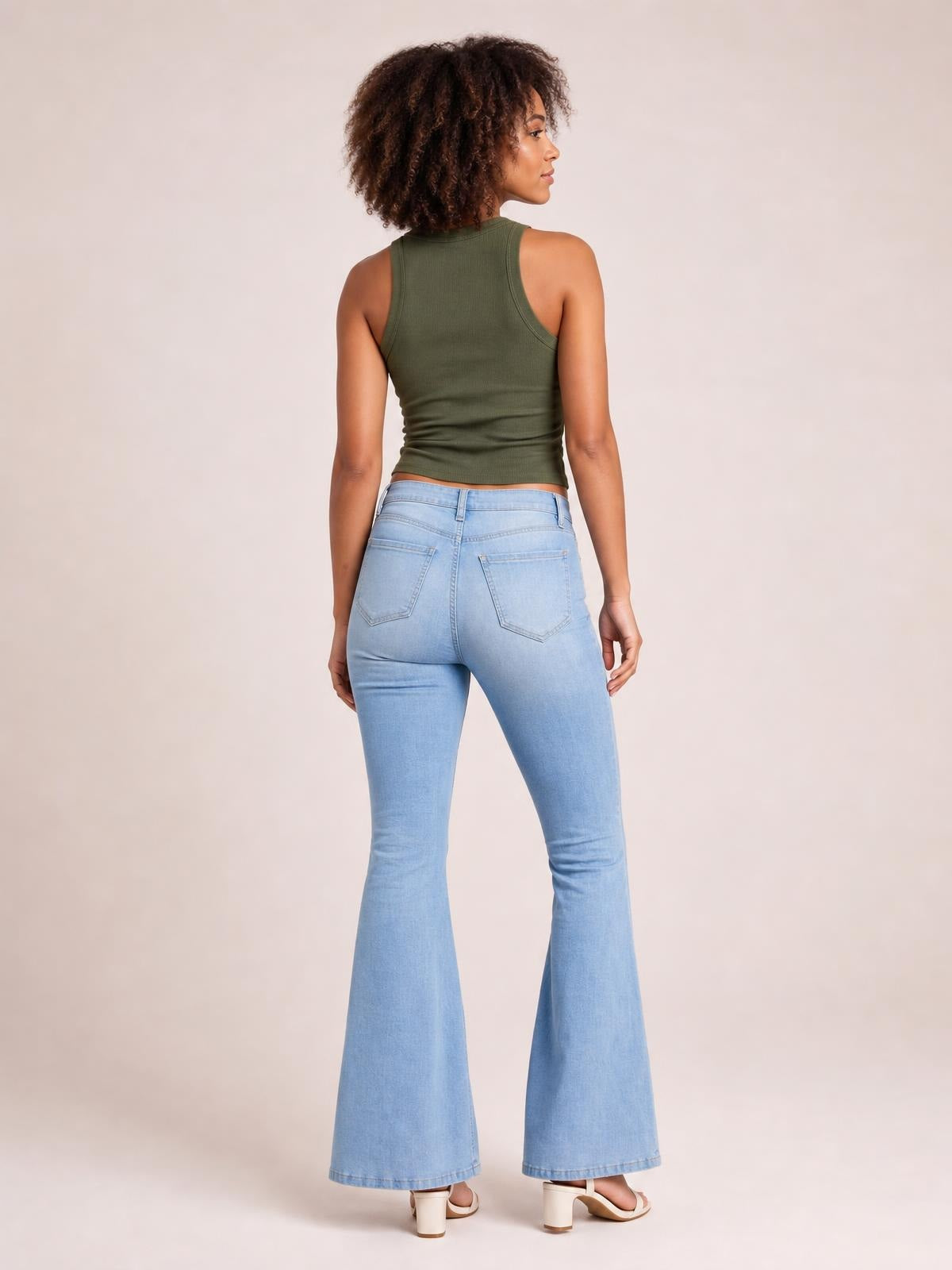 A model stands facing away in light blue jeans with stitched back pockets, a fitted seat, buttoned waistband, and wide flared legs.