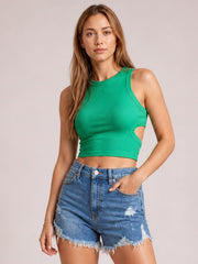 A woman modeling a bright green sleeveless crop top with side cutouts and light-wash distressed denim shorts against a solid light gray studio background.