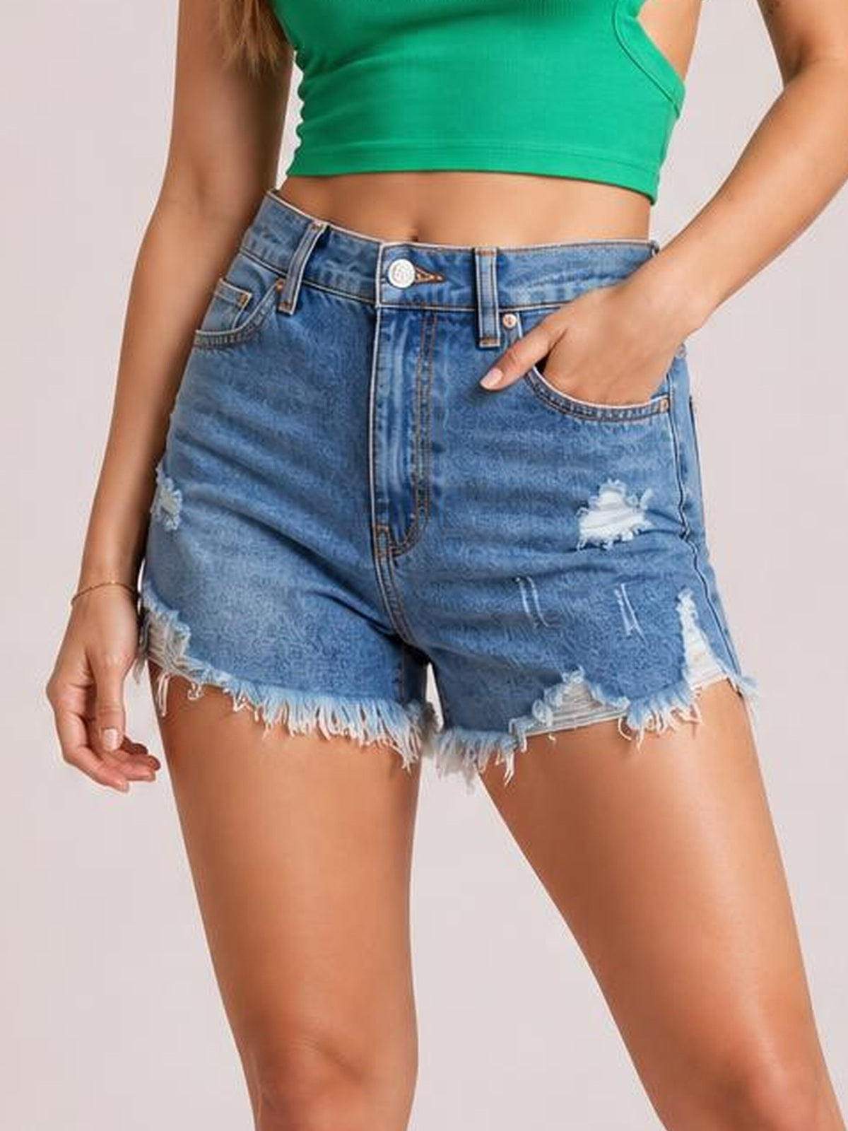 Model poses in blue distressed denim shorts with a high-rise silhouette, button fly, belt loops, pockets, and frayed raw hems.