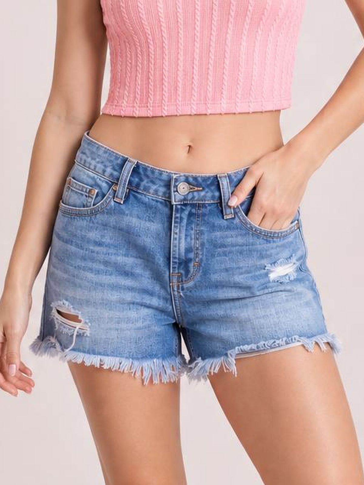 Model wears blue distressed denim shorts with a fitted silhouette, button fly, belt loops, front pockets, ripped details, and frayed raw hems, posing with one hand in a pocket.
