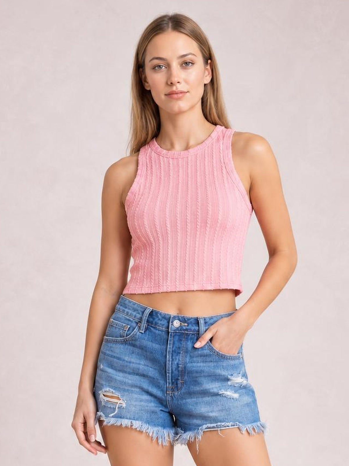 A woman modeling a pink ribbed knit sleeveless crop top paired with distressed blue denim shorts against a light beige studio background.