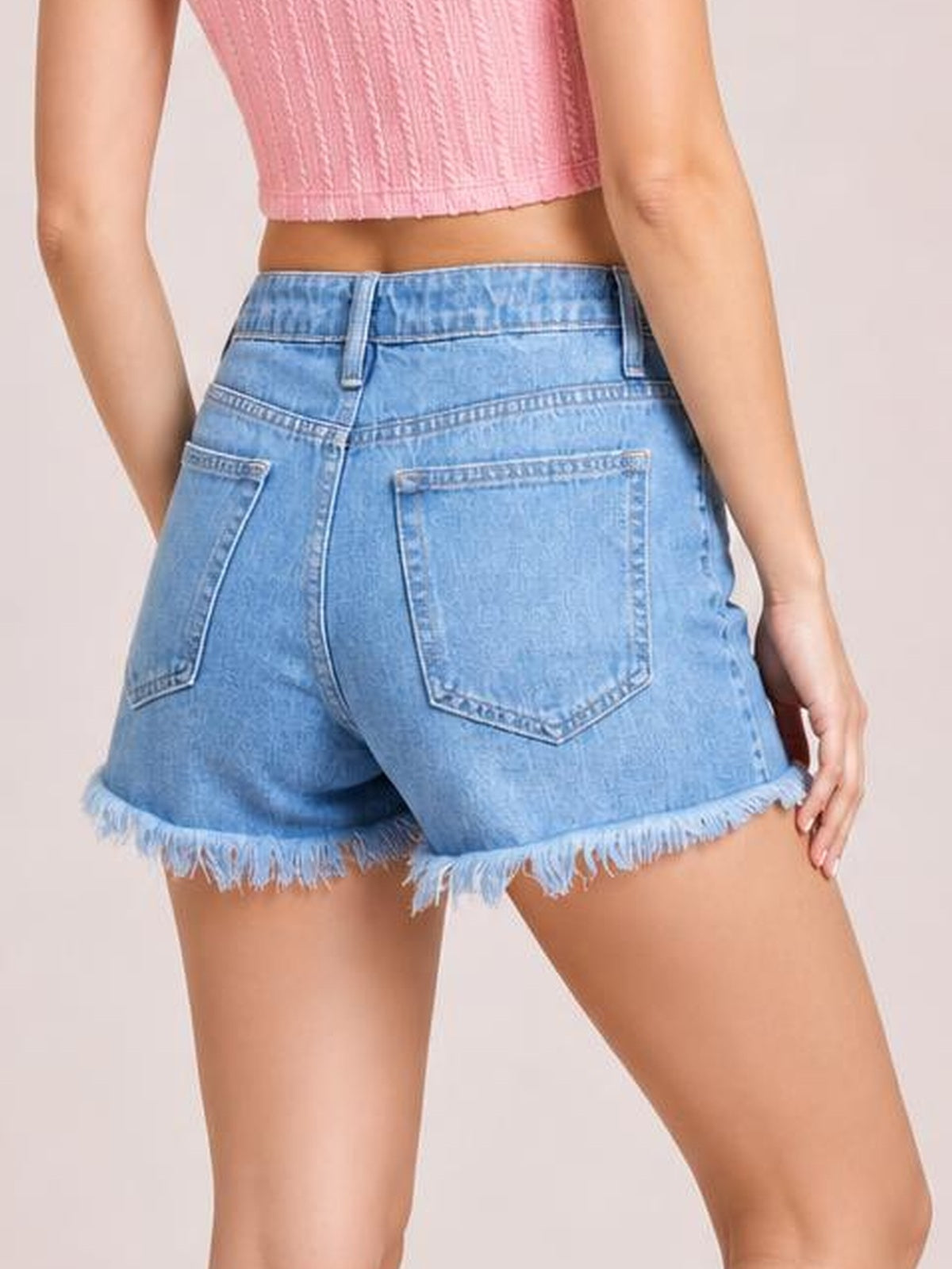 Model shown from behind wearing light blue distressed denim shorts with a high-rise waistband, belt loops, back patch pockets, and frayed raw hems.
