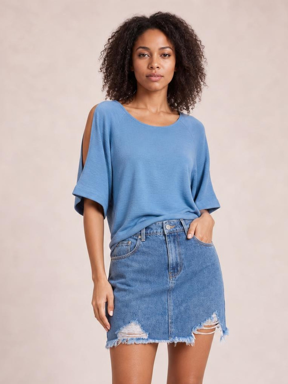 Model poses with one hand in the pocket of a blue distressed denim skirt featuring a front button closure, straight silhouette, ripped details, and raw frayed hem.