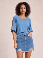 Model poses with one hand in the pocket of a blue distressed denim skirt featuring a front button closure, straight silhouette, ripped details, and raw frayed hem.