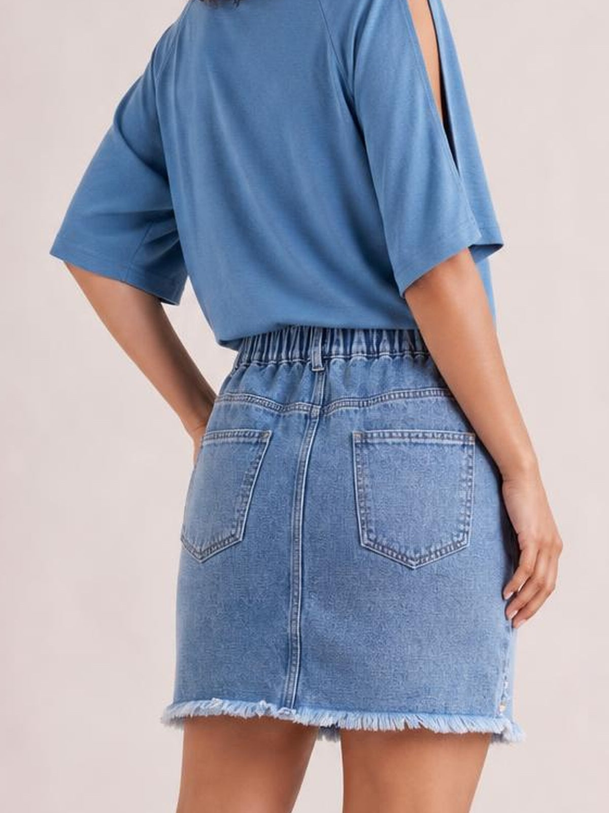 Model poses with one hand on her hip, showcasing a blue distressed denim skirt with a gathered waistband, back patch pockets, center seam, and raw hem.