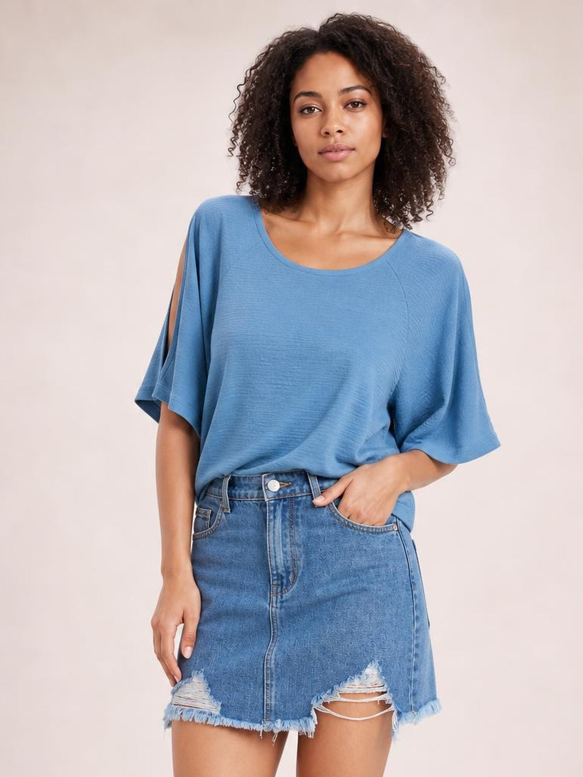 A model poses with one hand in the pocket of a blue denim skirt featuring a front button closure, distressed panels, pockets, and a frayed raw hem.