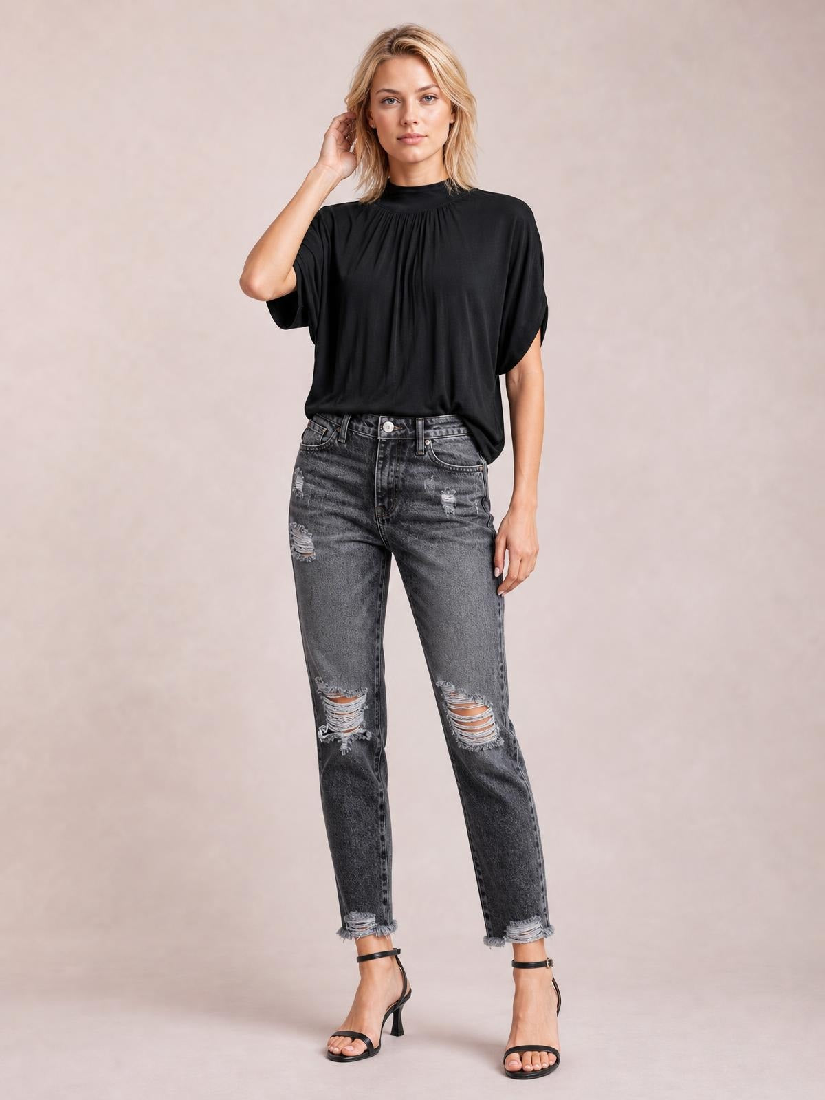 A model poses in dark gray distressed wash ankle jeans with a straight-leg silhouette, ripped knees, five-pocket construction, and raw hems.