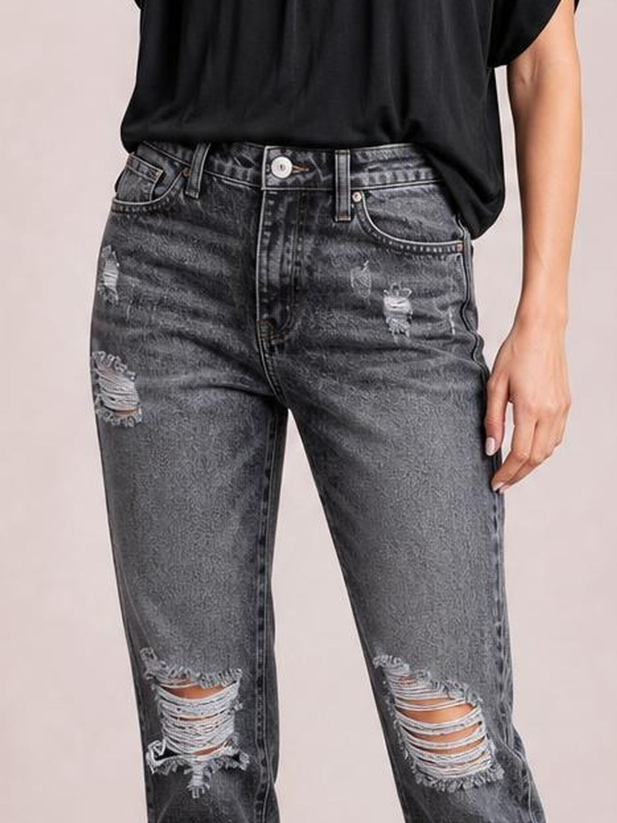 A model wears dark gray distressed wash jeans with a structured waistband, belt loops, five-pocket construction, and heavily ripped knees while standing with one hand at her side.