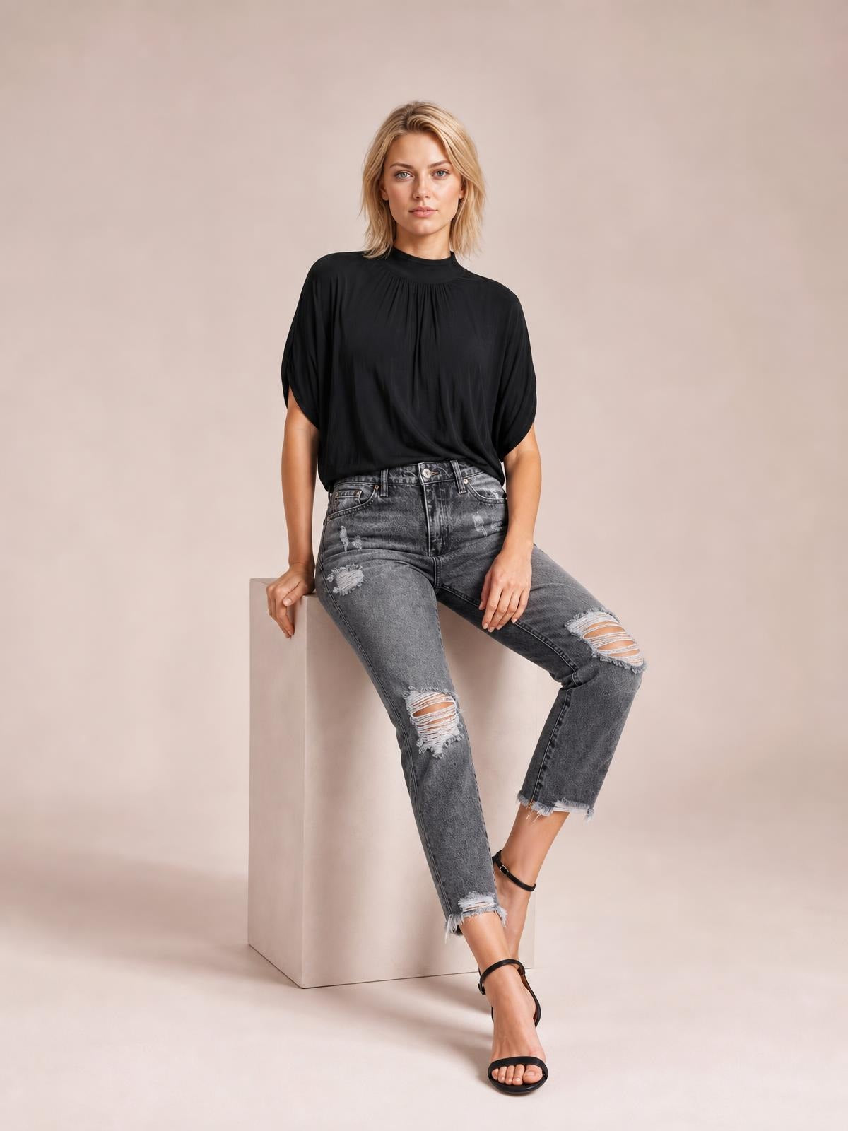 Model seated in distressed gray ankle jeans with a fitted straight-leg silhouette, multiple ripped details, front pockets, belt loops, and raw hems.