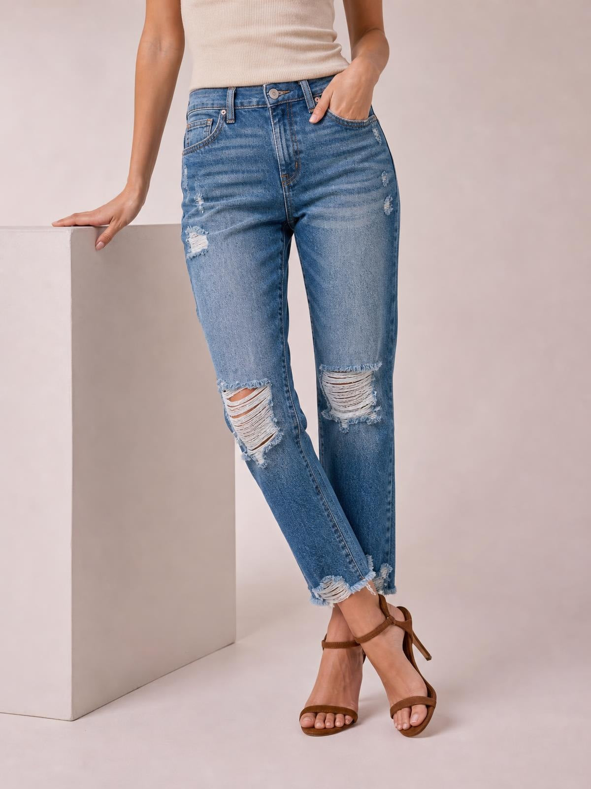 A model poses in blue distressed wash ankle jeans with a straight-leg silhouette, five-pocket construction, ripped knees, and frayed raw hems.