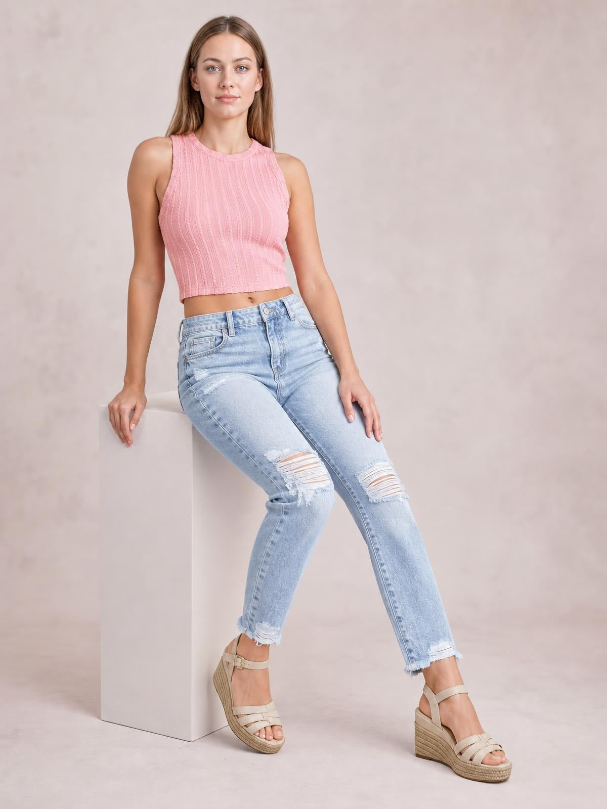 A model sits with one leg extended, wearing light-wash distressed ankle jeans with a slim silhouette, ripped knees, belt loops, and raw hems.