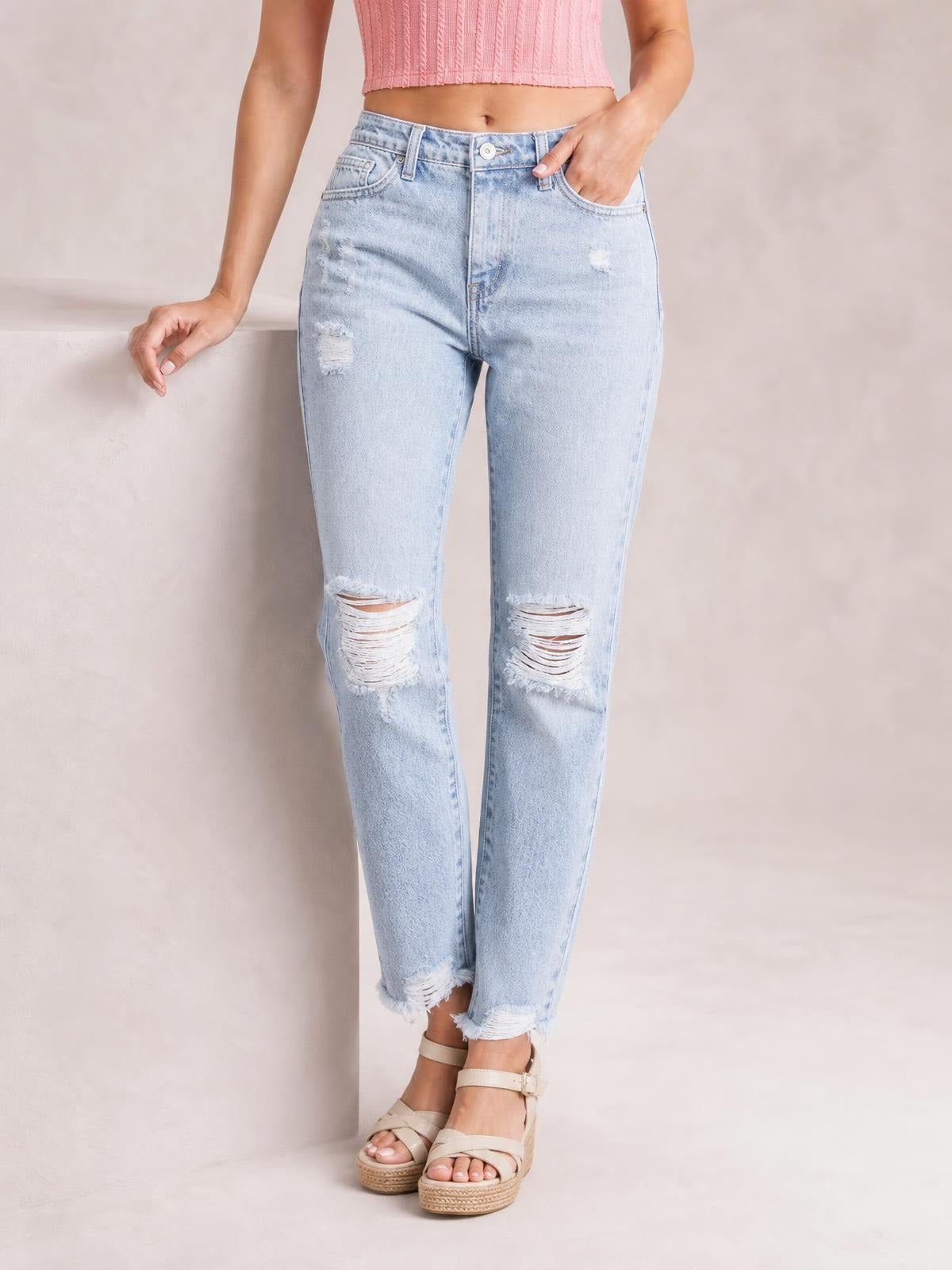 Model poses in light-wash distressed ankle jeans with a straight silhouette, ripped knees, five-pocket construction, belt loops, and raw hems.