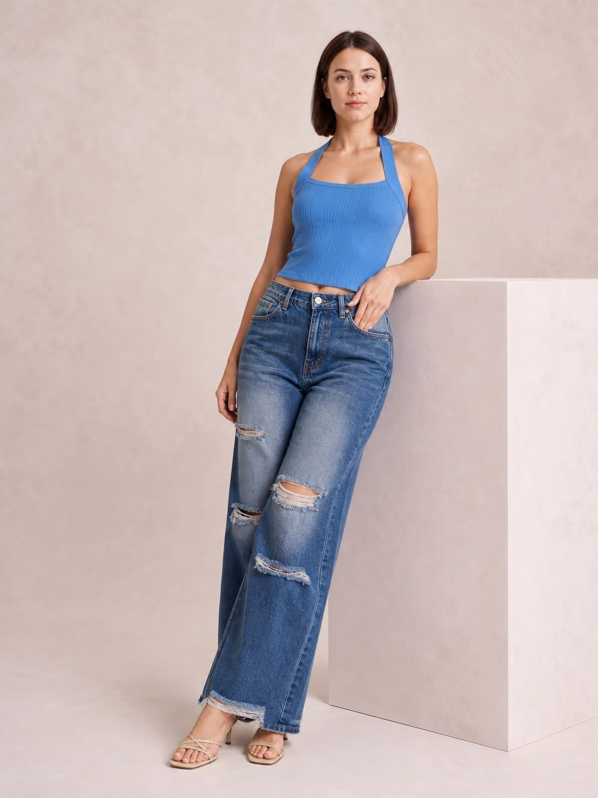 A model poses in blue distressed wide leg jeans with a button fly, front pockets, relaxed silhouette, ripped knees, and frayed hems.