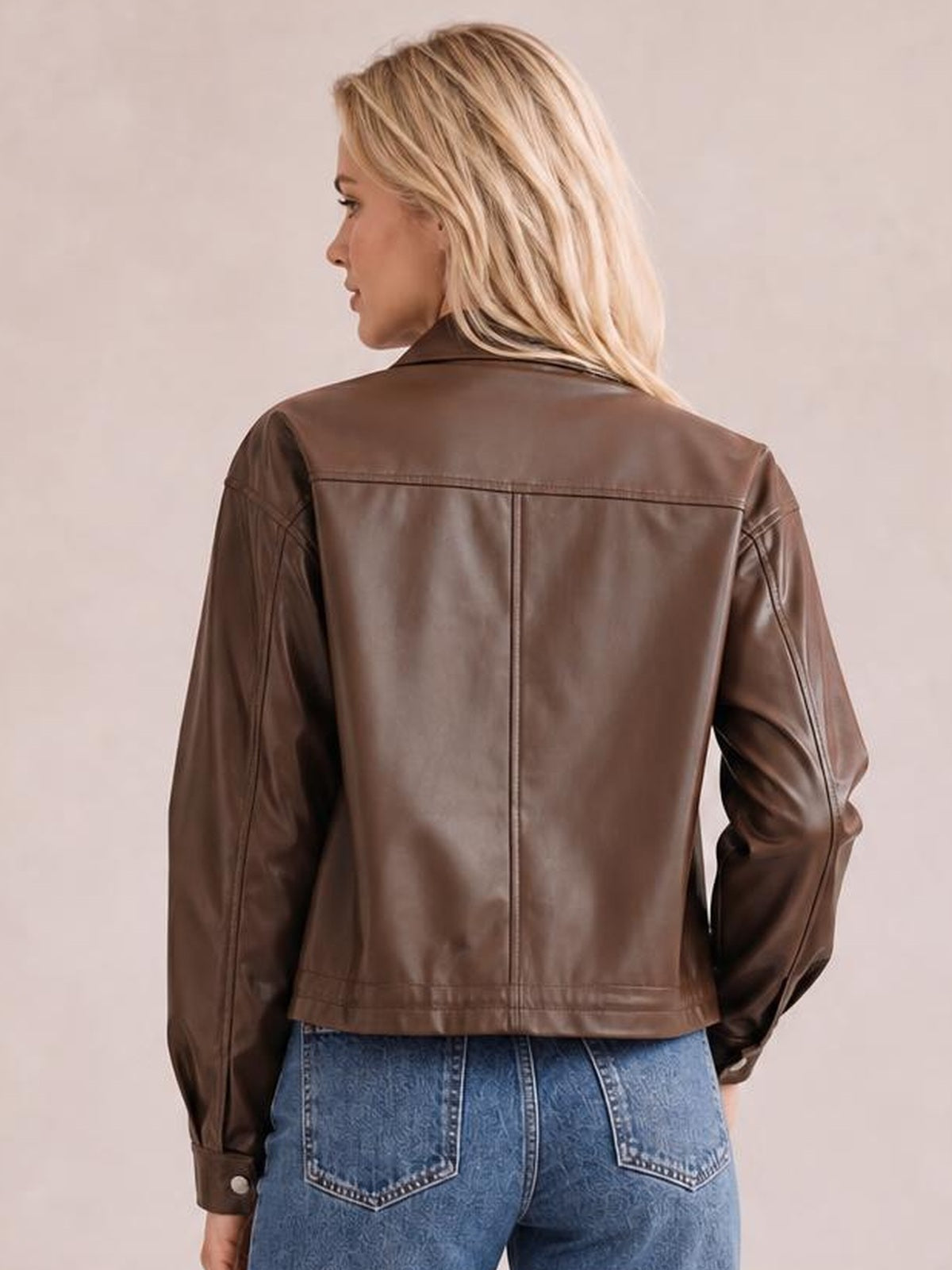 A model poses in profile wearing a brown leather jacket with a structured collar, long sleeves, seamed back construction, and a straight drawstring hem silhouette.