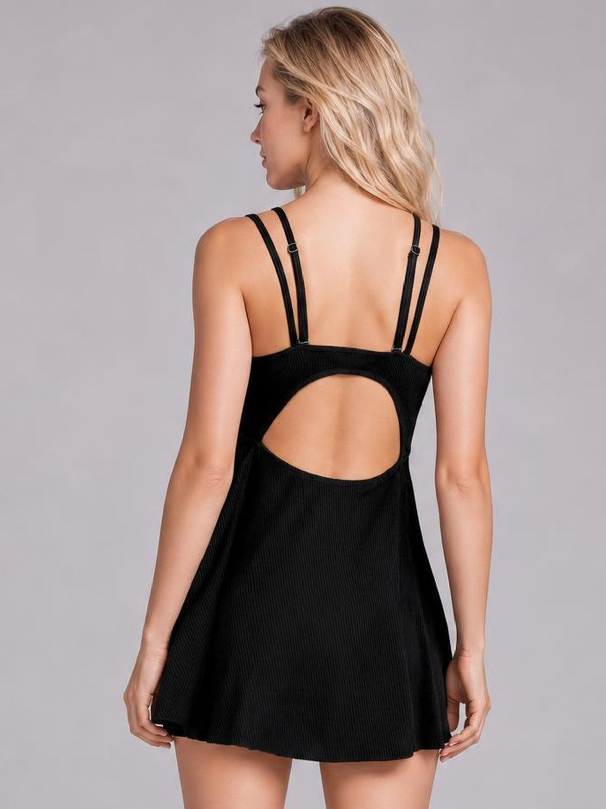 A model faces away in a black double-strap cutout swim dress with adjustable straps, an oval open back, and a flared skirt silhouette.
