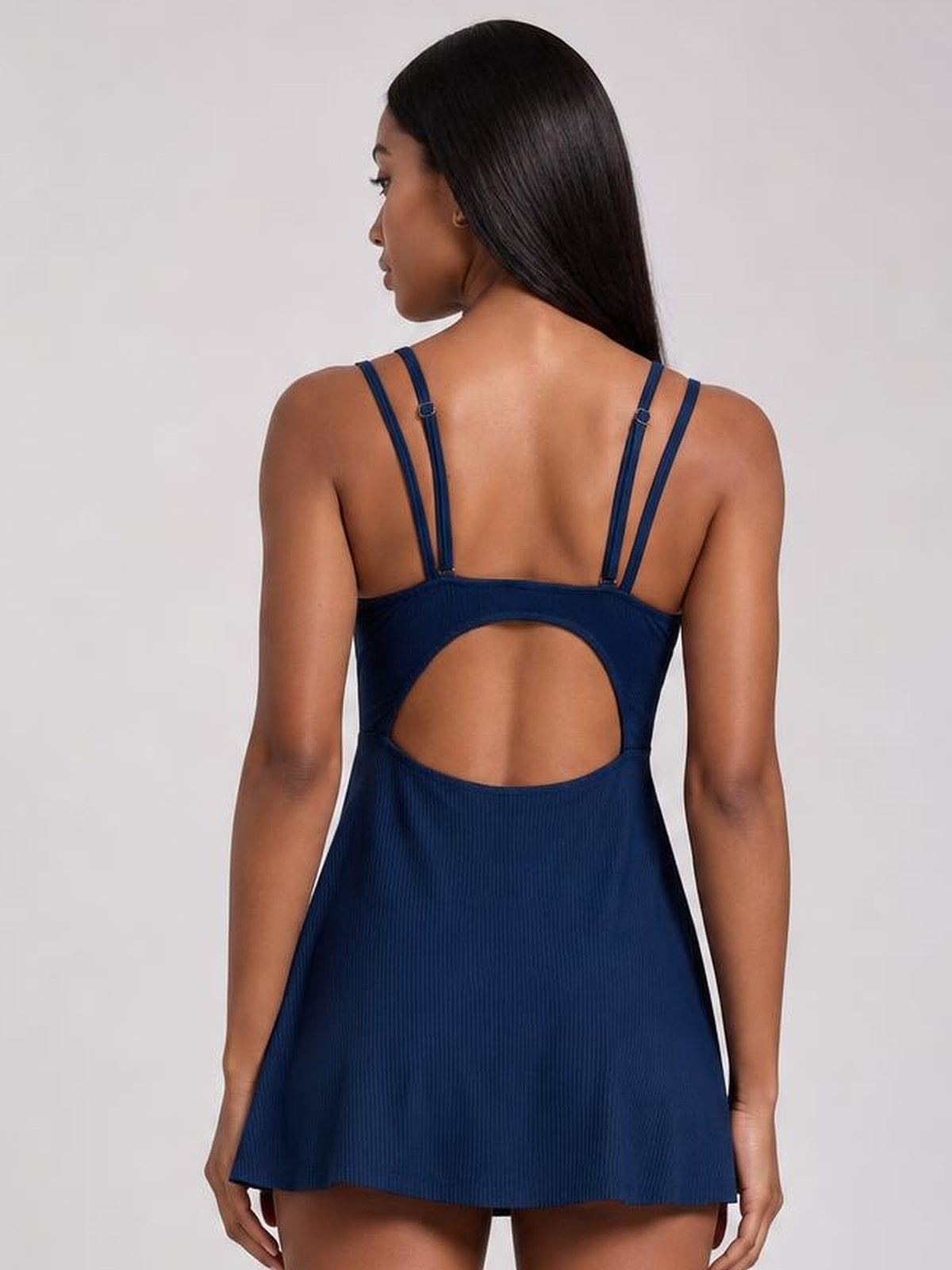 Back view of a model standing in a solid navy double-strap cutout swim dress with adjustable straps, a large oval back cutout, and a flared skirt silhouette.