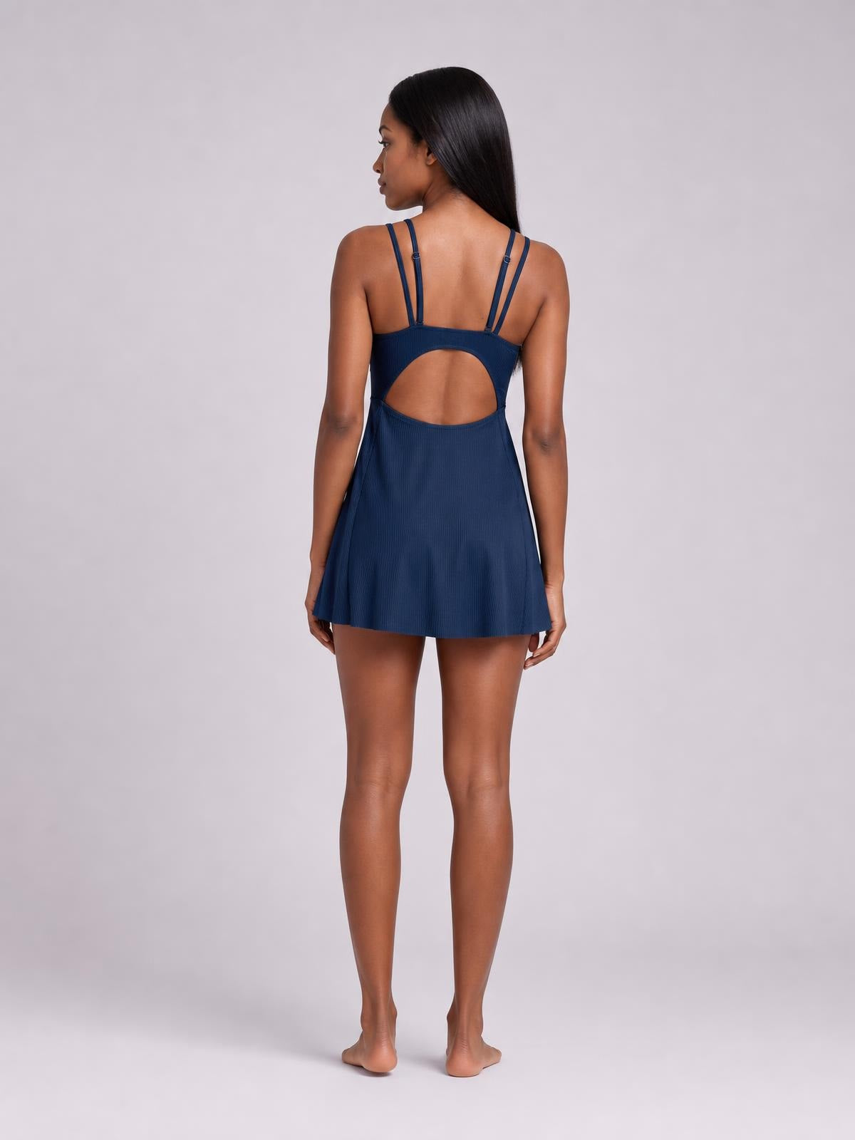 Model stands facing away in a solid navy Double-Strap Cutout Swim Dress with twin shoulder straps, a large oval back cutout, and a flared skirt silhouette.