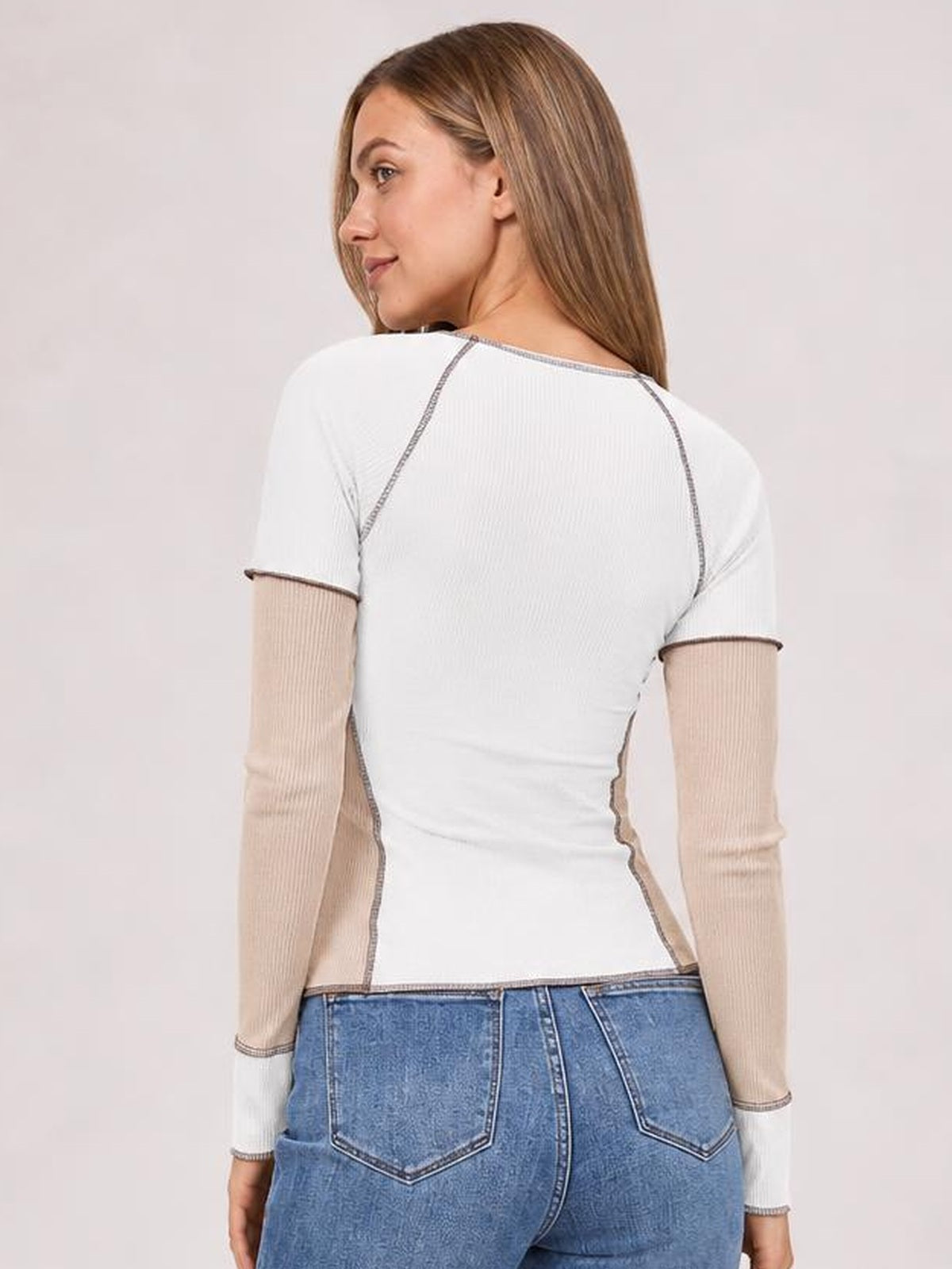 Model looks over her shoulder wearing a fitted white ribbed color block top with beige long sleeves, short sleeve overlays, and exposed contrast seams.