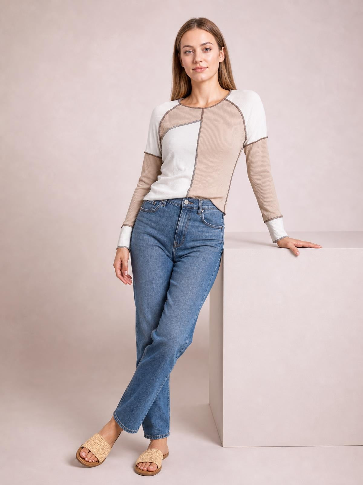 Model wears a fitted color block top in white and beige with long ribbed sleeves, exposed contrast seams, and a curved hem while leaning with crossed legs.